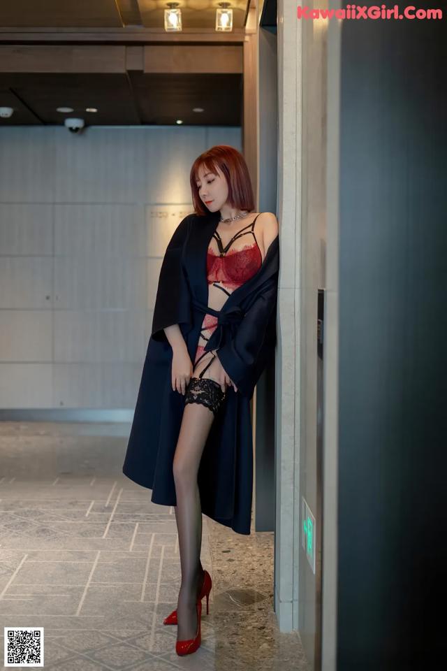 A woman in a black robe and red lingerie leaning against a wall.