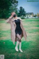 A woman in a black dress and a beige trench coat is standing in the grass.